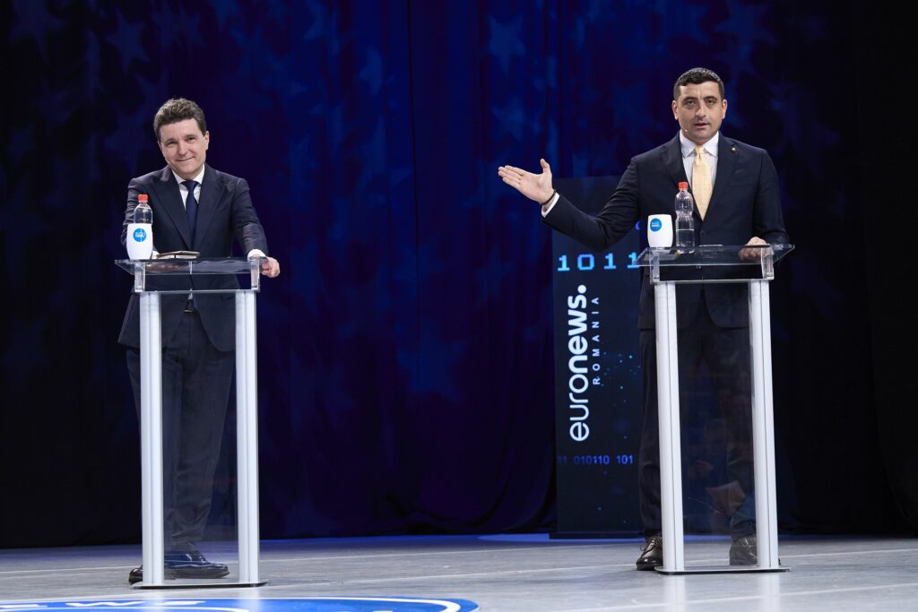 Two political candidates in suits standing behind podiums. the one on the right, Simon, gestures to the one on the left (Dan) the stage is white, background is dark blue, almost black